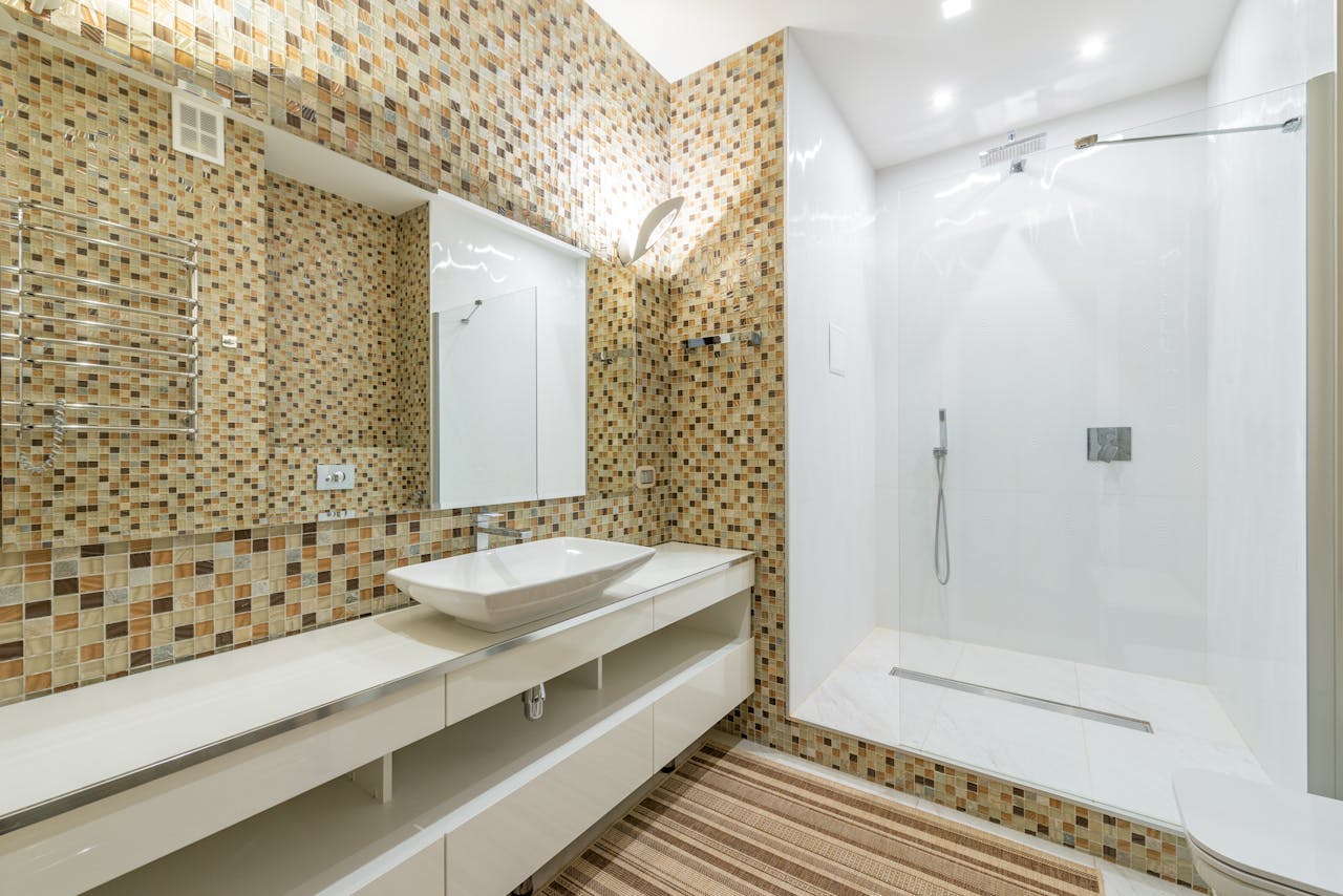 Wide angle of interior of contemporary bathroom with shower cabin and large mirror above white clean sink at mosaic tiled wall